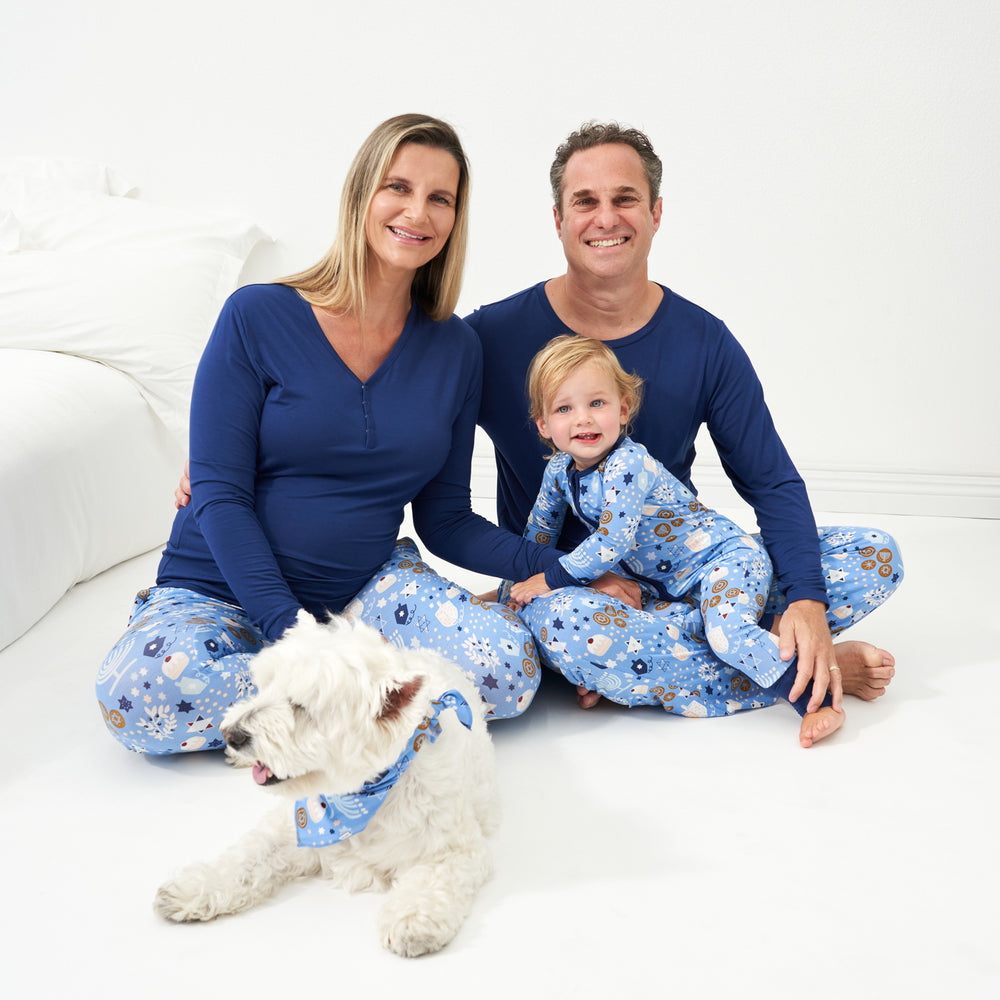 Family of three posing together wearing Hanukkah Lights and Love pajama sets. Dad is wearing a Hanukkah Lights and Love men's pajama pants and Sapphire men's top. Mom is wearing Hanukkah Lights and Love women's pajama pants and Sapphire women's pajama top. Child is wearing a Hanukkah Lights and Love zippy and their dog is wearing a Hanukkah Lights and Love pet bandana