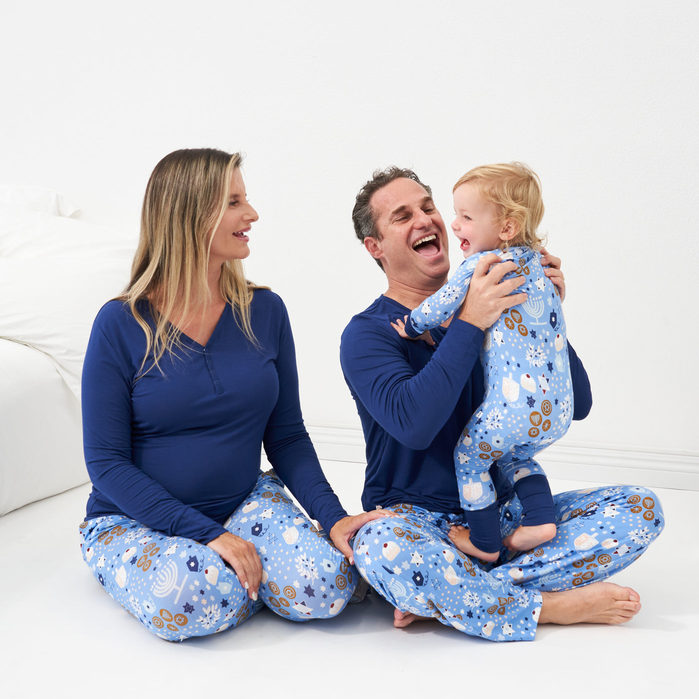 Family of three posing together wearing Hanukkah Lights and Love pajama sets. Dad is wearing a Hanukkah Lights and Love men's pajama pants and Sapphire men's top. Mom is wearing Hanukkah Lights and Love women's pajama pants and Sapphire women's pajama top. Child is wearing a Hanukkah Lights and Love zippy. 