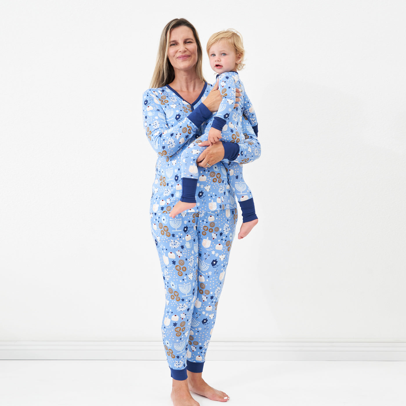 Woman holding her child on her hip wearing Hanukkah Lights and Love women's pajama top paired with matching women's pajama bottoms. Her child is wearing a Hanukkah Lights and Love zippy