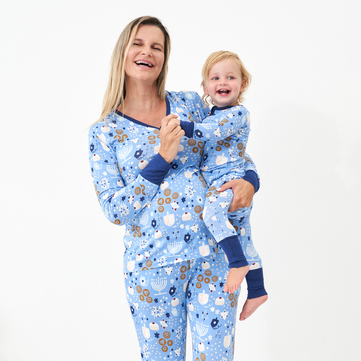 Woman holding her child on her hip wearing Hanukkah Lights and Love women's pajama top paired with matching women's pajama bottoms. Her child is wearing a Hanukkah Lights and Love zippy