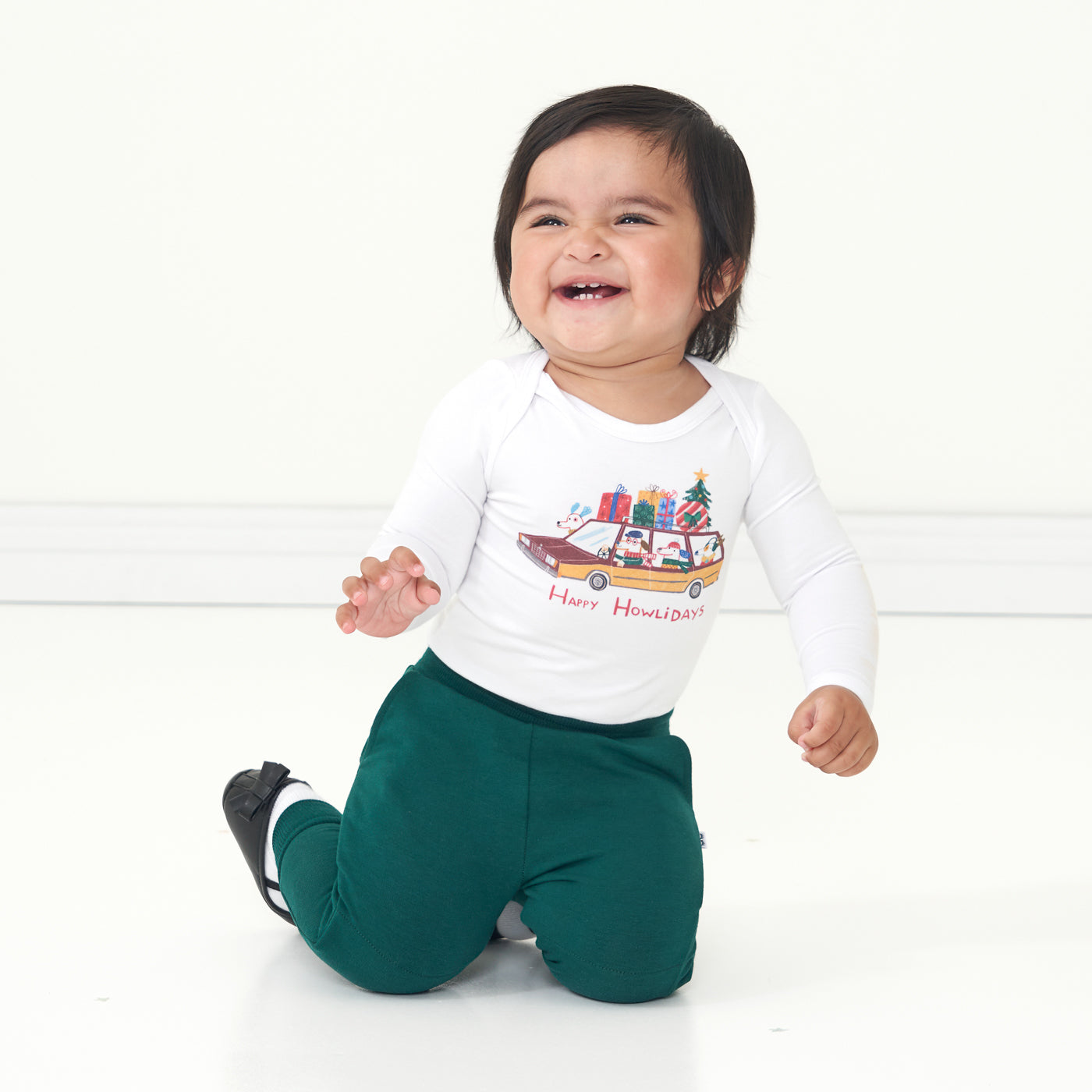 Child kneeling on the ground wearing a Happy Howlidays graphic bodysuit and coordinating emerald joggers