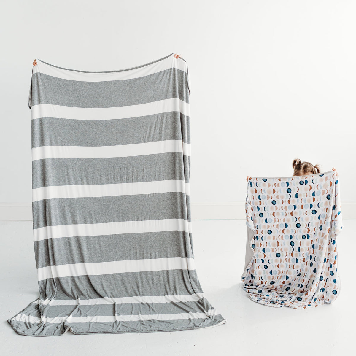 Mother and child holding up bamboo blankets. Mom is holding up a Heather Gray Wide Stripe oversized bamboo blanket. Child is holding up a Luna Neutral large bamboo blanket