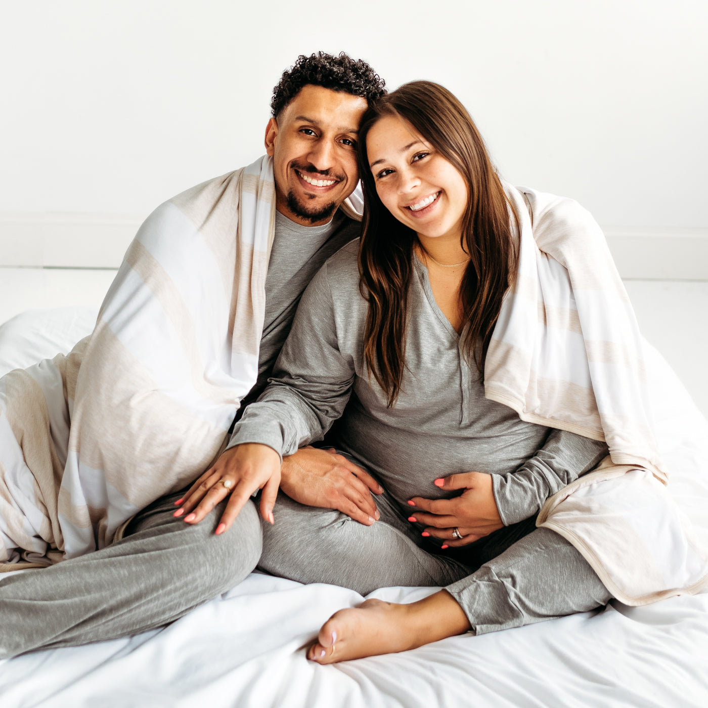Couple sitting on the ground, wearing matching pajamas, with a Heather Oatmeal Stripe oversized bamboo blanket wrapped around them.