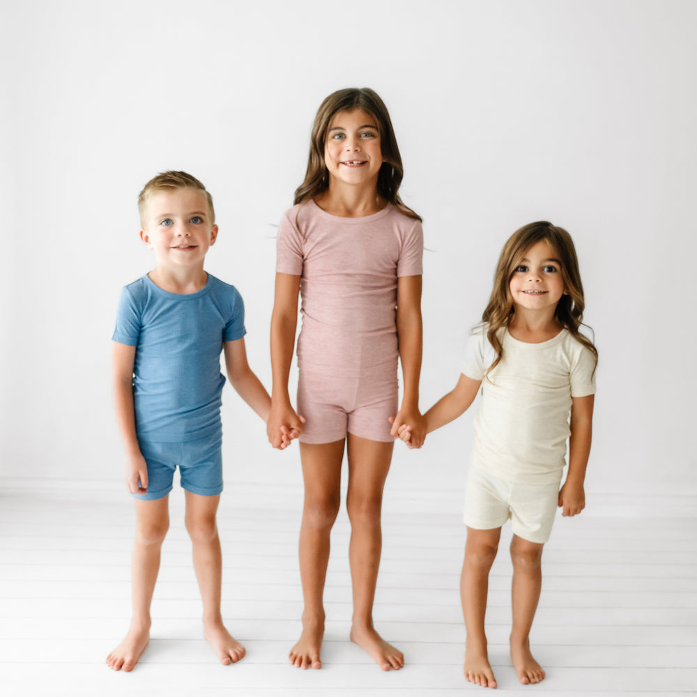 Three children holding hands wearing coordinating Ribbed two piece short sleeve and shorts pajama sets in Heather Blue, Heather Mauve, and Heather Oatmeal