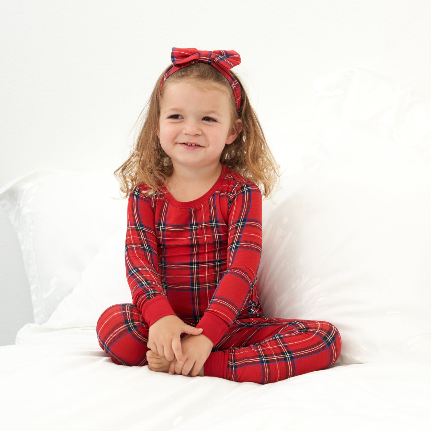 Child sitting on a bed wearing a Holiday Plaid two piece pajama set and matching luxe bow headband