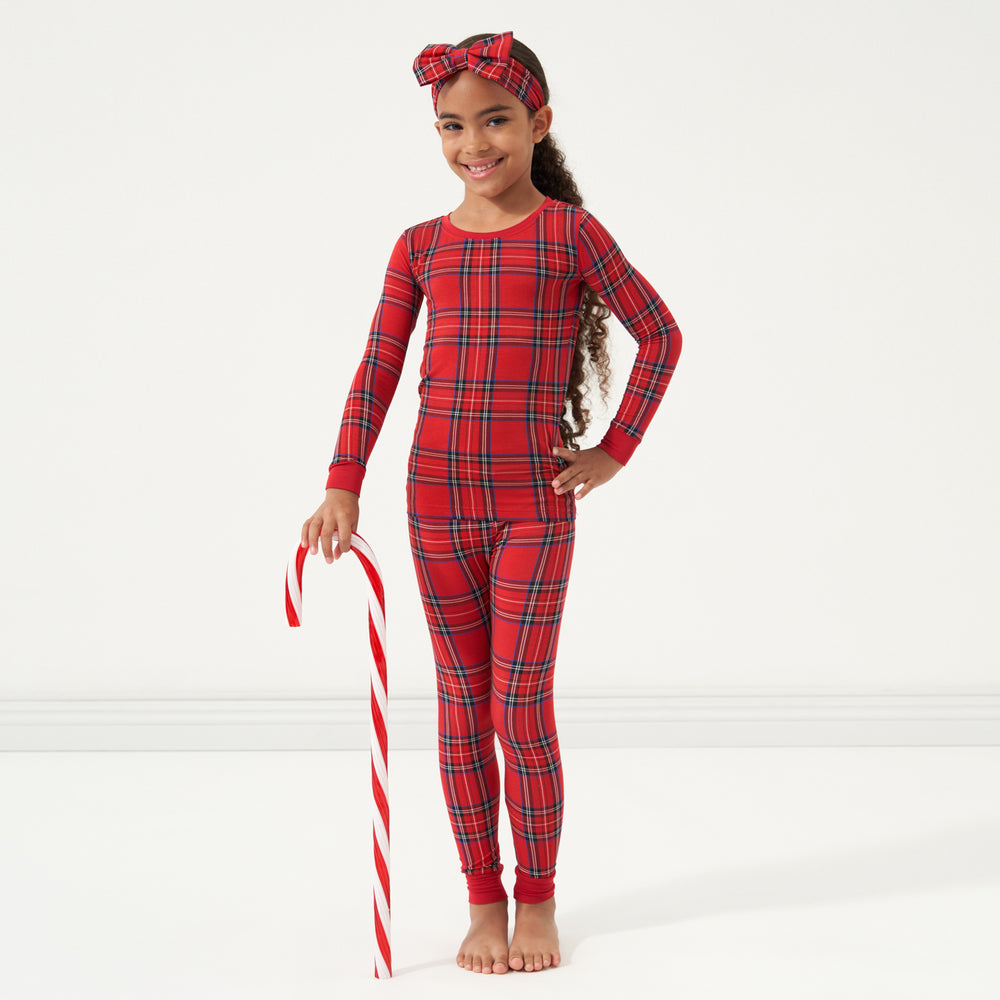 Child wearing a Holiday Plaid two piece pajama set and matching luxe bow headband holding a large candy cane