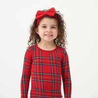 Close up image of a child wearing a Holiday Plaid two piece pajama set and coordinating Holiday Red luxe bow headband