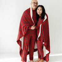 Man and woman wearing matching Holiday Plaid men's and women's pajama sets wrapped in a Holiday Plaid oversized plush blanket