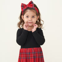 Child with her hands on her face wearing a Holiday Plaid skater dress with bodysuit and matching luxe bow headband