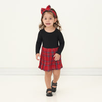 Child walking forward wearing a Holiday Plaid skater dress with bodysuit and matching luxe bow headband