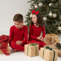 Two children sitting in front of a tree with presents wearing matching Holiday Red two-piece pajama sets