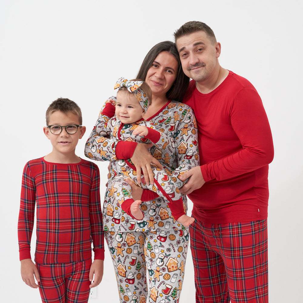 Family of four posing together. Mom is wearing a women's Holiday Treats pajama top paired with women's Holiday Treats pajama pants. Dad is wearing men's Holiday Red pajama top paired with men's Holiday Plaid pajama pants. One child is wearing a Holiday Plaid two piece pajama set and his sibling is wearing a Holiday Treats zippy paired with a matching Holiday Treats luxe bow headband.