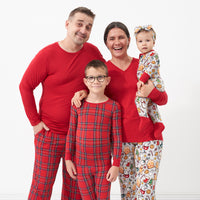 Family of four posing together. Mom is wearing a women's Holiday Red pajama top paired with women's Holiday Treats pajama pants. Dad is wearing men's Holiday Red pajama top paired with men's Holiday Plaid pajama pants. One child is wearing a Holiday Plaid two piece pajama set and his sibling is wearing a Holiday Treats zippy paired with a matching Holiday Treats luxe bow headband.