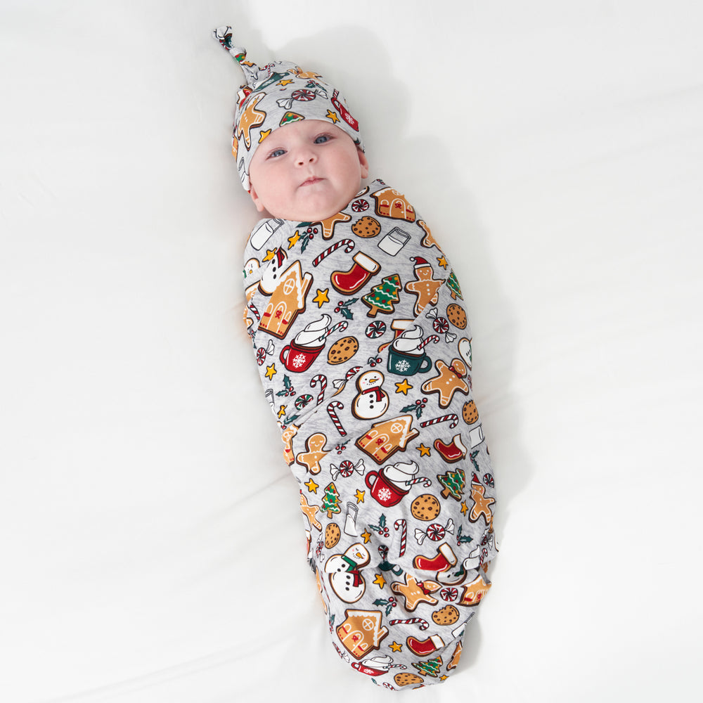 Alternate image of a child laying on a bed swaddled in a Holiday Treats swaddle and hat set