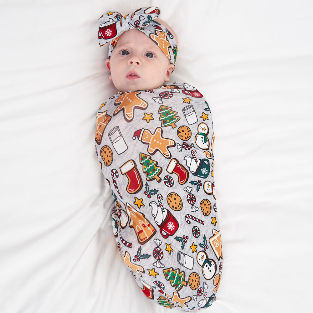 Child laying on a bed swaddled in a Holiday Treats swaddle and luxe bow headband set