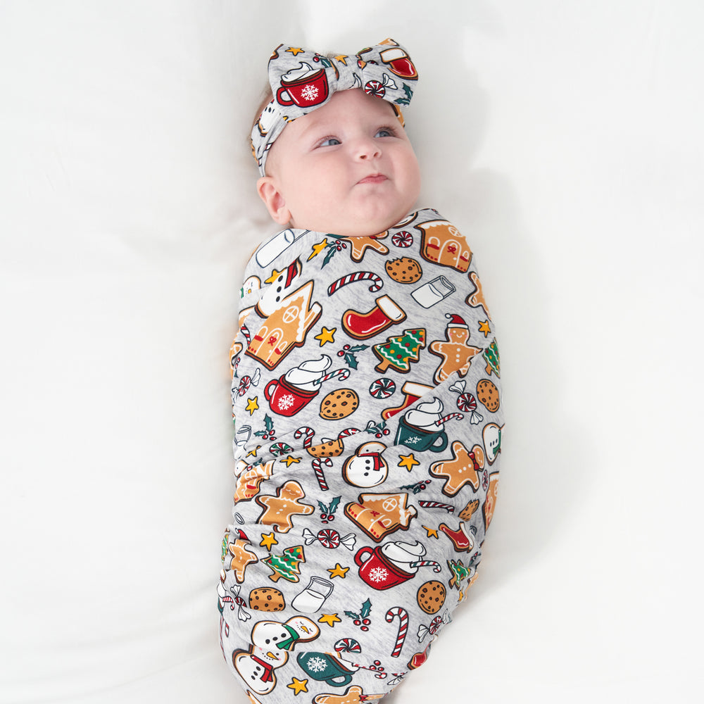 Alternate image of a child laying on a bed swaddled in a Holiday Treats swaddle and luxe bow headband set
