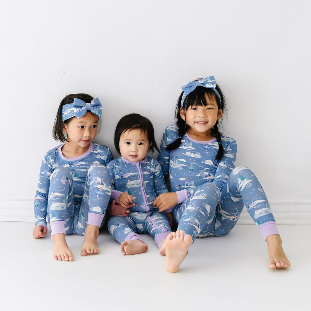 Three children sitting together wearing matching Ice Princess pajamas in two piece and zippy styles. Two children have a matching Ice Princess luxe bow paired with their pjs.