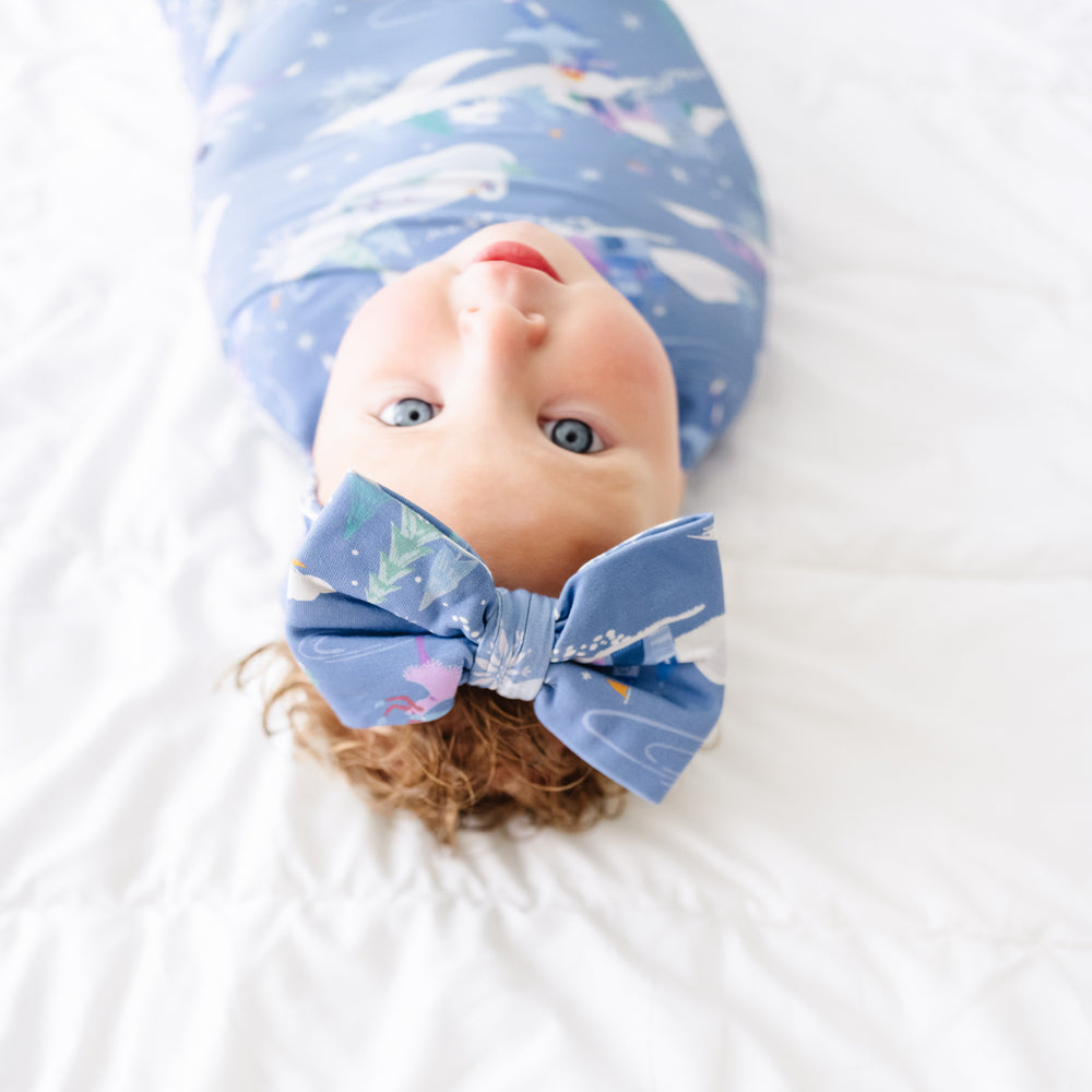 Close up image of a child laying on a bed swaddled in an Ice Princess printed swaddle and luxe bow headband set