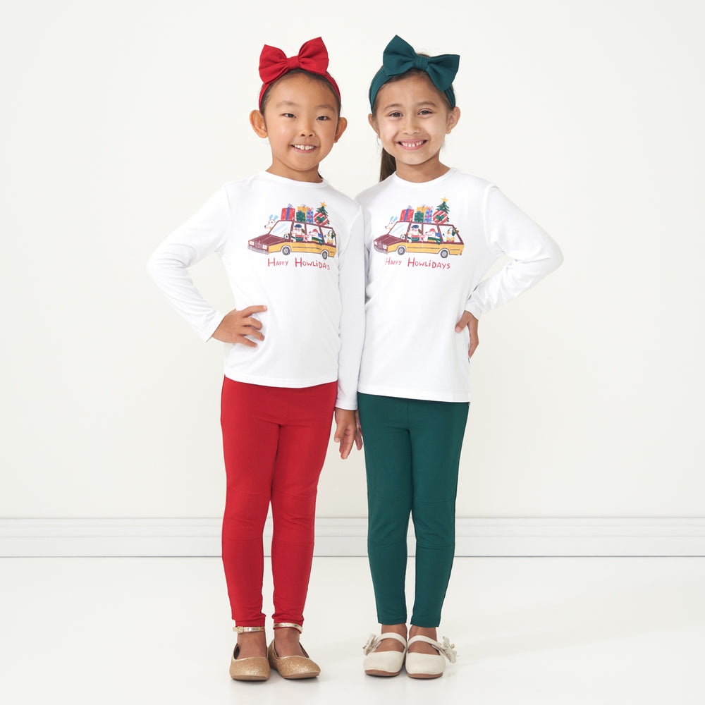 Two children posing together wearing matching Happy Howlidays graphic tees. Each child is pairing their graphic tee with Emerald and Holiday Red leggings and coordinating Emerald and Holiday Red luxe bow headbands. 