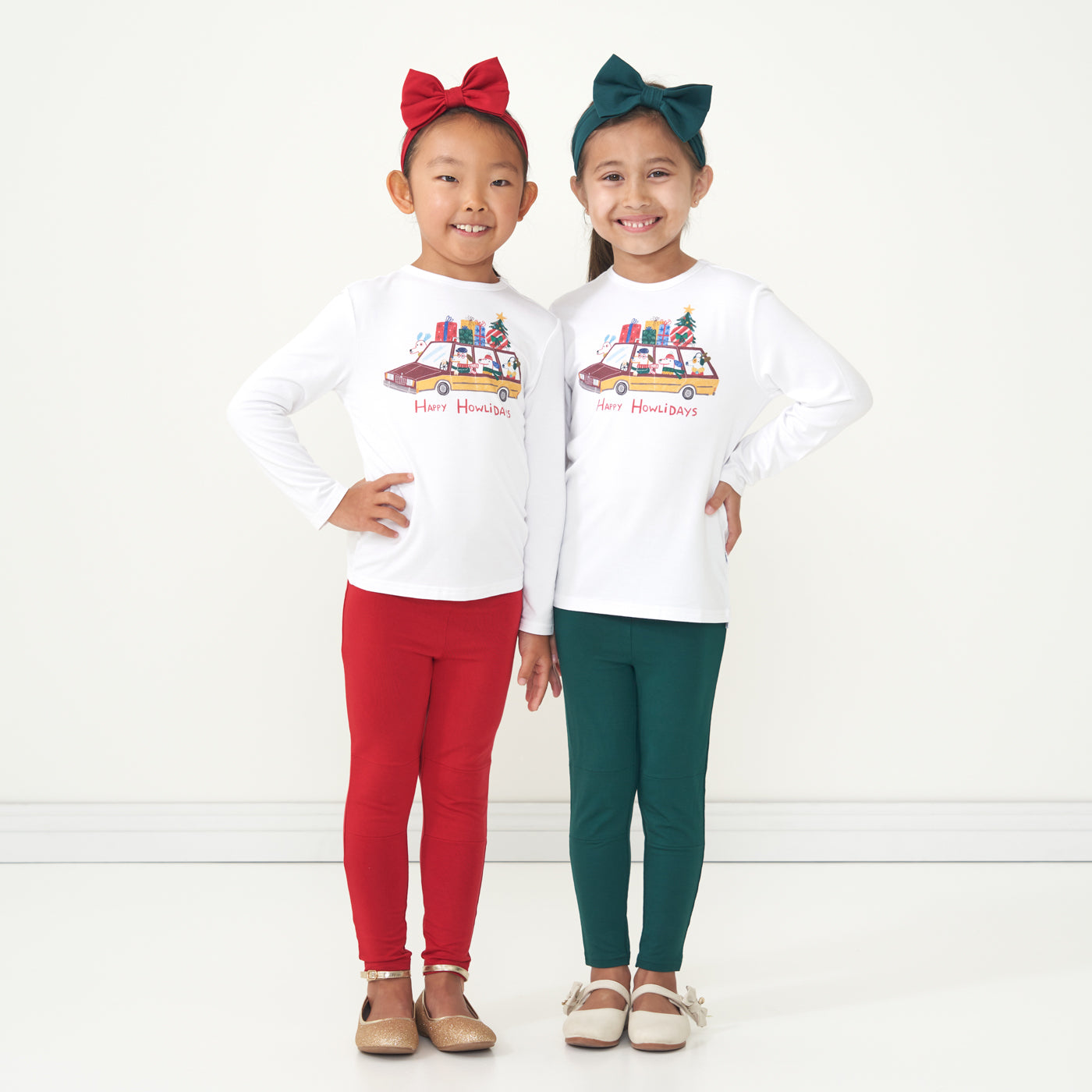 Two children posing together wearing matching Happy Howlidays graphic tees. Each child is pairing their graphic tee with Emerald and Holiday Red leggings and coordinating Emerald and Holiday Red luxe bow headbands. 
