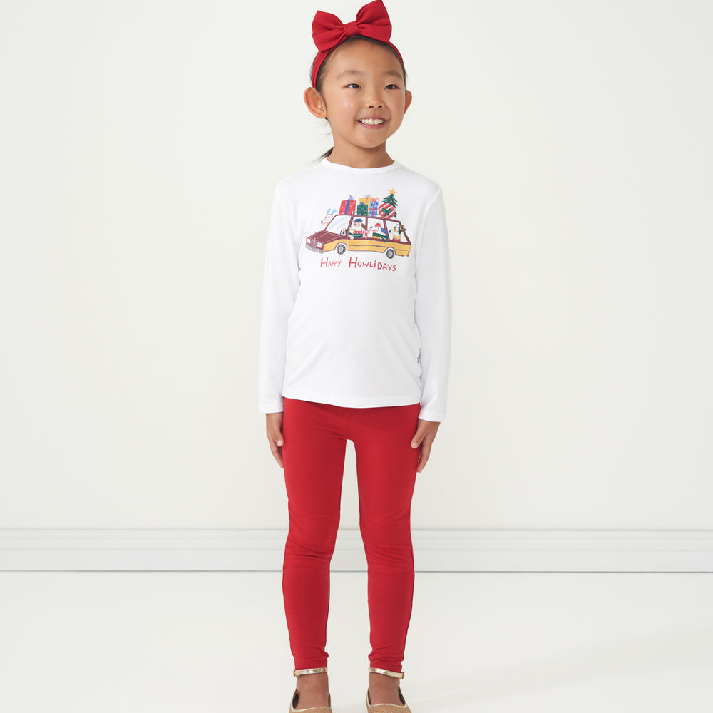 Child posing wearing Holiday Red leggings paired with a Happy Howlidays graphic tee and matching Holiday Red luxe bow headband