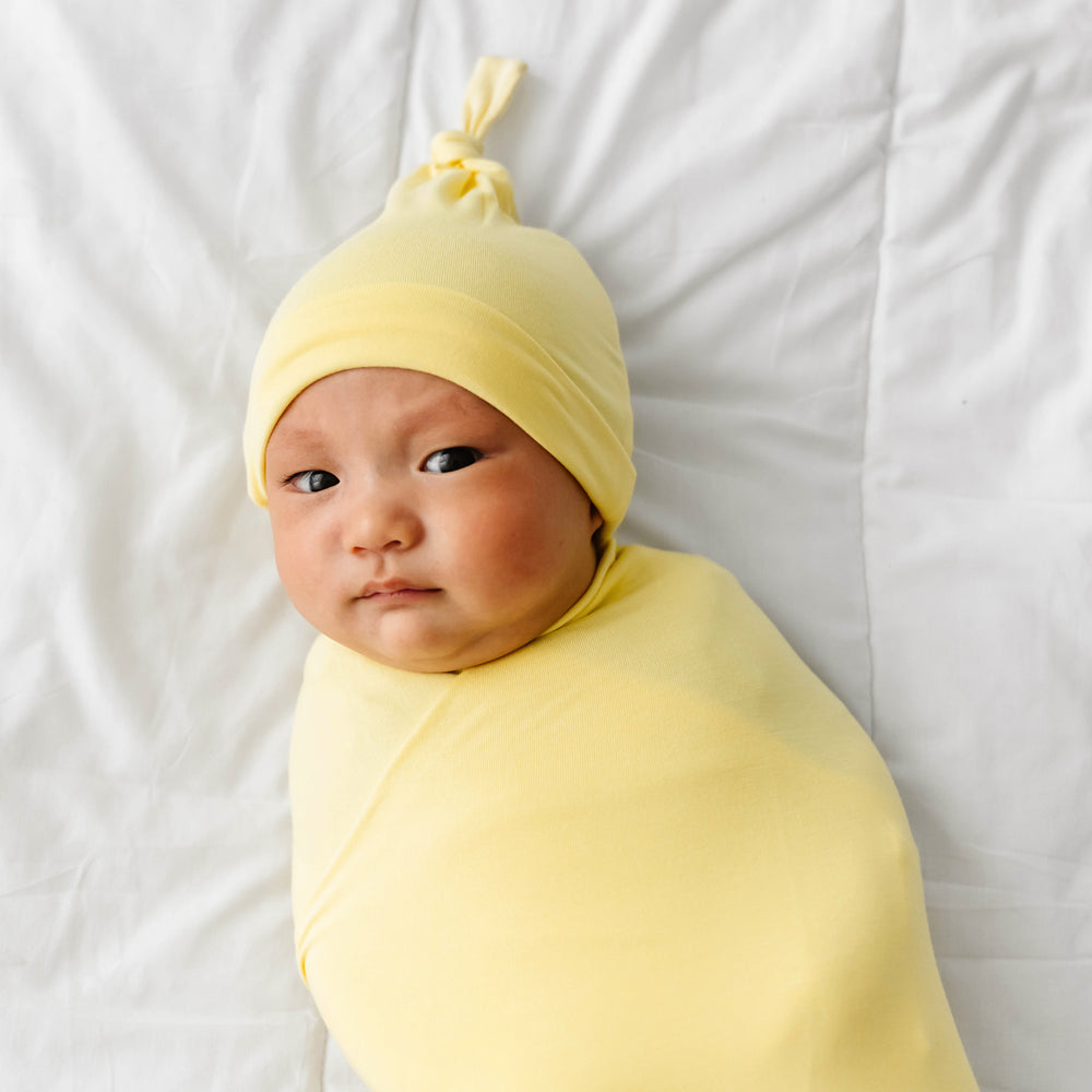 Close up image of a child laying on a bed swaddled in a Lemon Twist swaddle and hat set