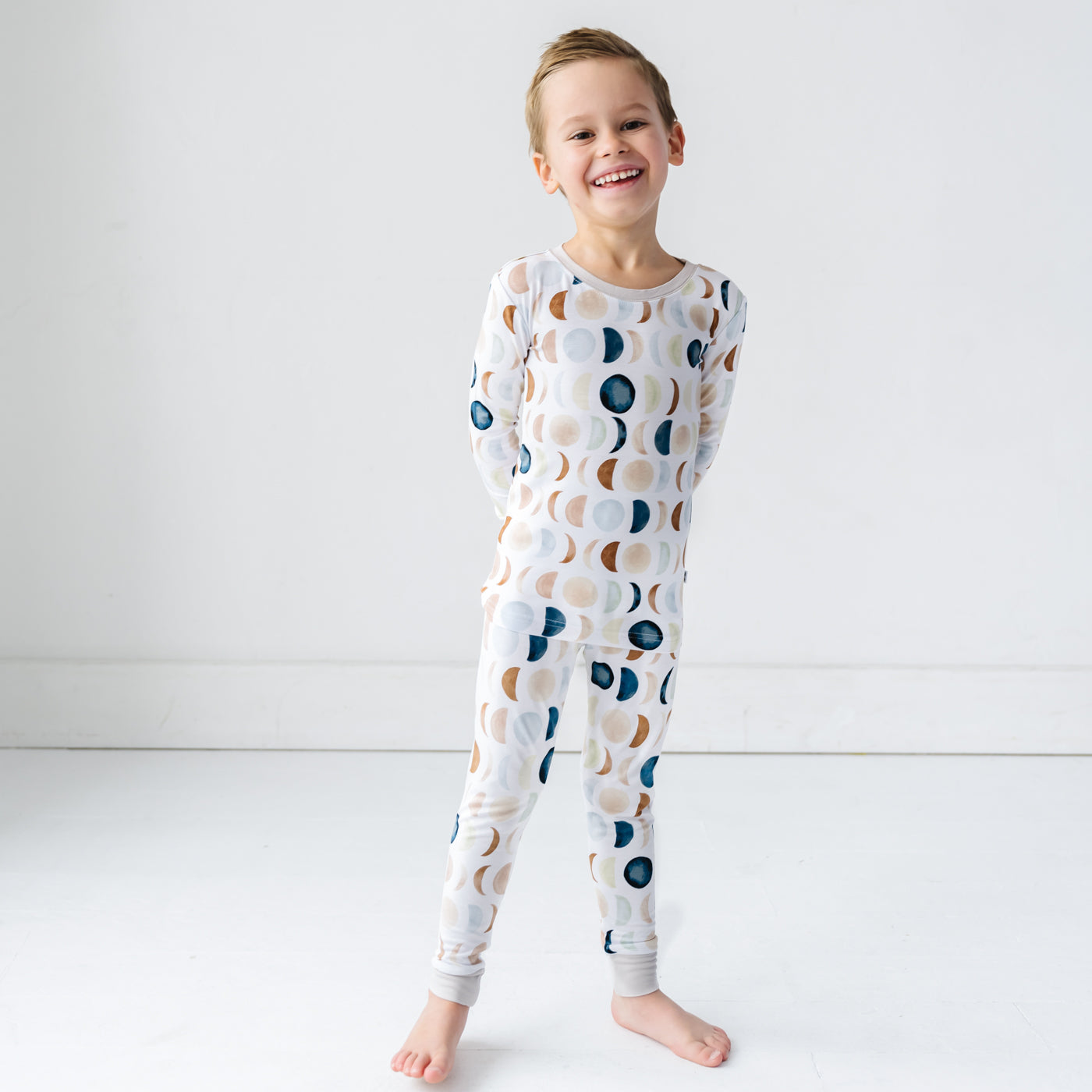 Image of a toddler in long sleeve top and pajama pants set in Luna Neutral print. This print features phases of the moon in the sweetest shades of creams, tans, and navy watercolor in an all over repeat pattern.