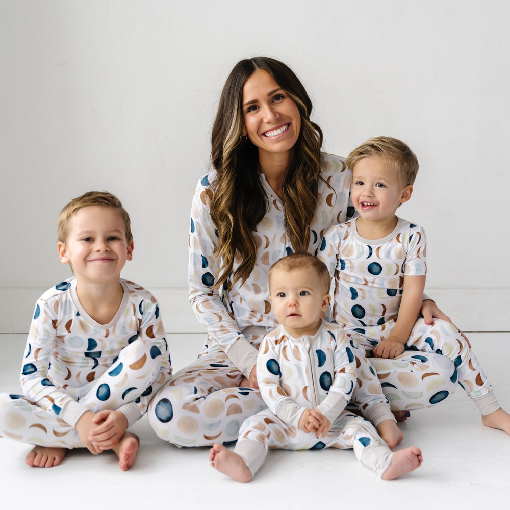 Mother and her three children sitting together wearing matching Luna Neutral pajamas. Mom is wearing women's Luna Neutral pajama top paired with matching pajama pants. Children are wearing Luna Neutral two piece and zippy style pjs