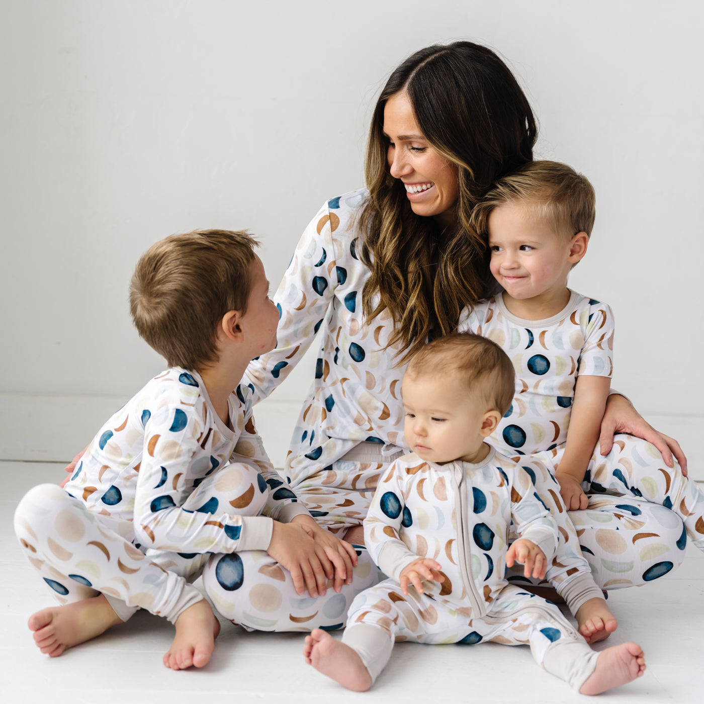 Image of a mother and three young children in matching pajama sets in Luna Neutral print. This print features phases of the moon in the sweetest shades of creams, tans, and navy watercolor in an all over repeat pattern.