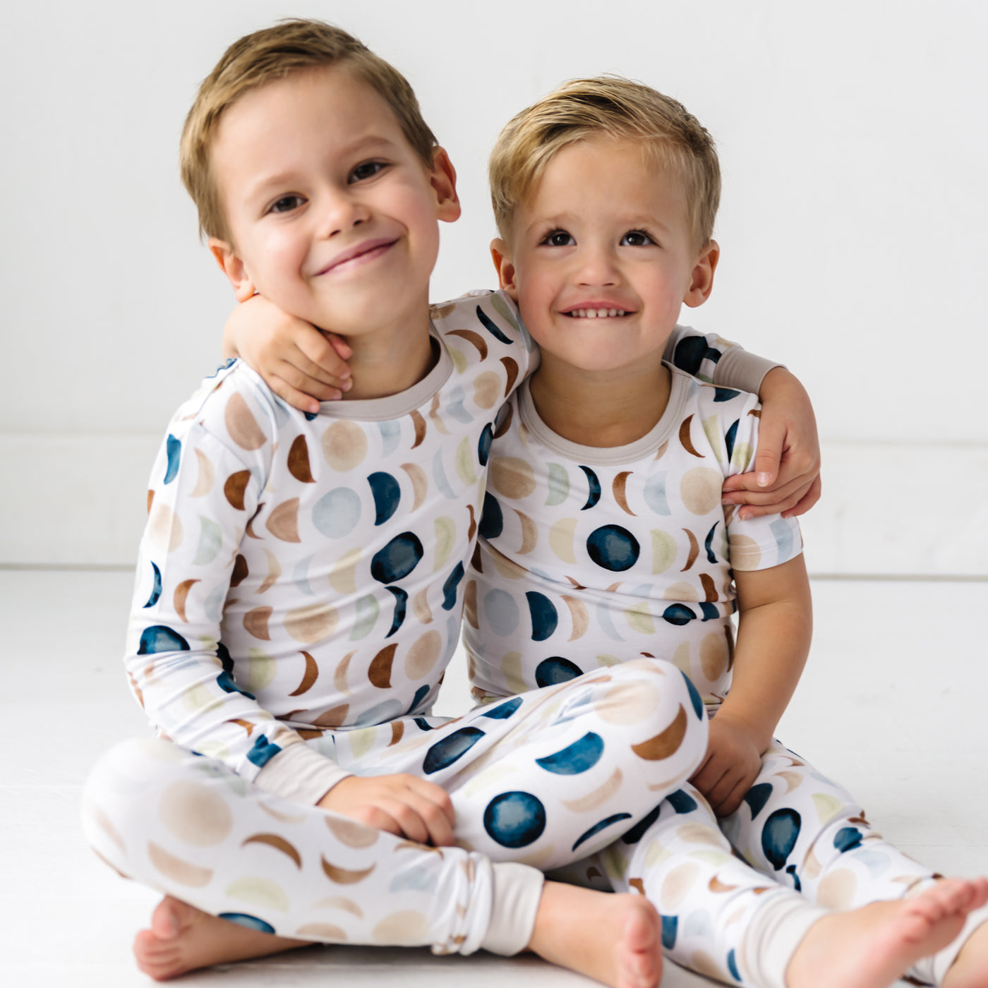 Image of two toddlers in pajama sets in Luna Neutral print. This print features phases of the moon in the sweetest shades of creams, tans, and navy watercolor in an all over repeat pattern.