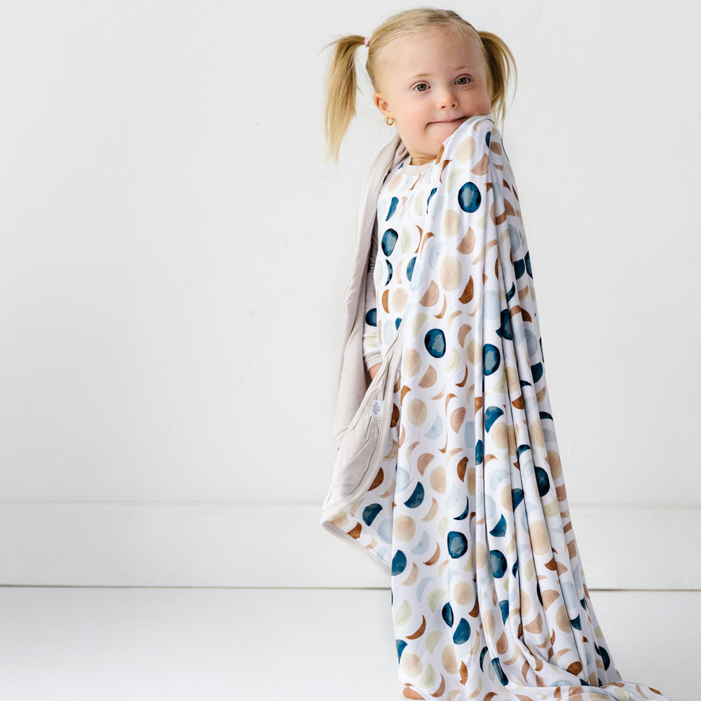 Image of a toddler girl with a triple layer bamboo viscose blanket draped around her shoulders in Luna Neutral print. This print features phases of the moon in the sweetest shades of creams, tans, and navy watercolor in an all over repeat pattern.