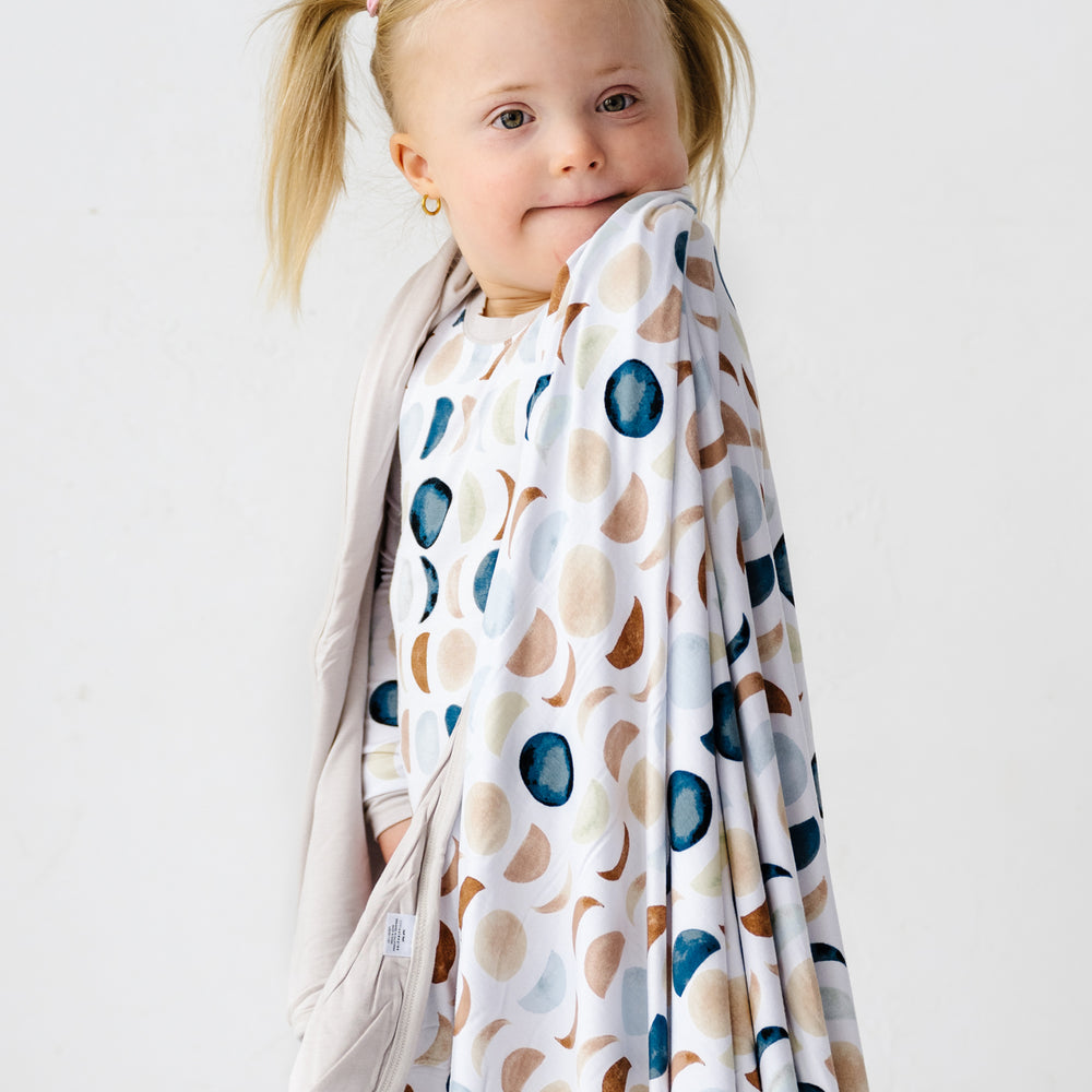 Close up image of a toddler girl with a triple layer bamboo viscose blanket draped around her shoulders in Luna Neutral print. This print features phases of the moon in the sweetest shades of creams, tans, and navy watercolor in an all over repeat pattern.
