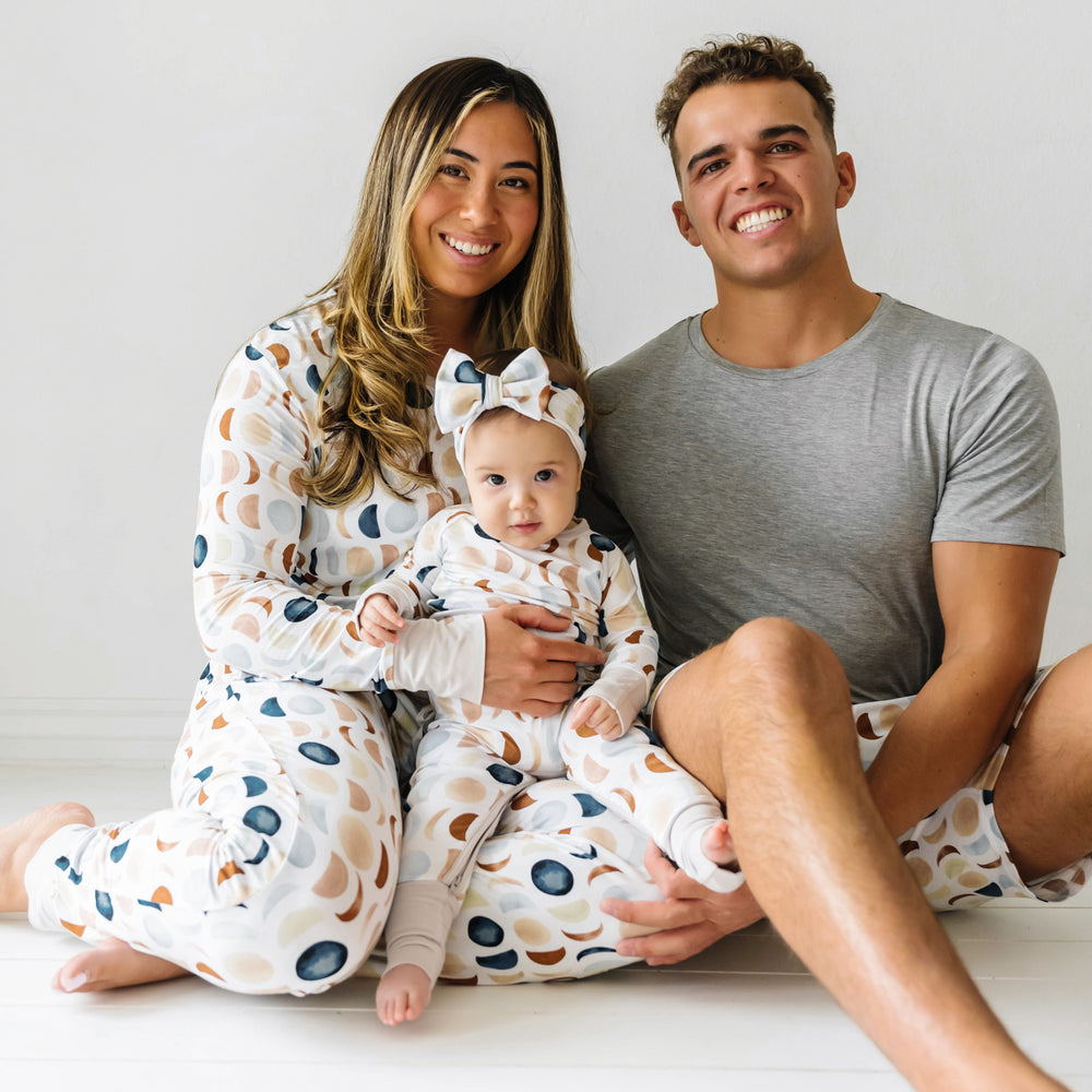 Click to see full screen - Family of three wearing coordinating Luna Neutral pajamas. Mom is wearing a Luna Neutral women's pajama top paired with matching women's pants. Dad is wearing a Heather Gray men's short sleeve pj top paired with Luna Neutral men's pj shorts. Child is wearing a Luna Neutral crescent zippy paired with a matching luxe bow headband.