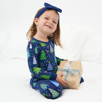 Child sitting on a bed holding a Christmas present wearing a Blue Merry and Bright two piece pajama set paired with a Sapphire Luxe Bow headband