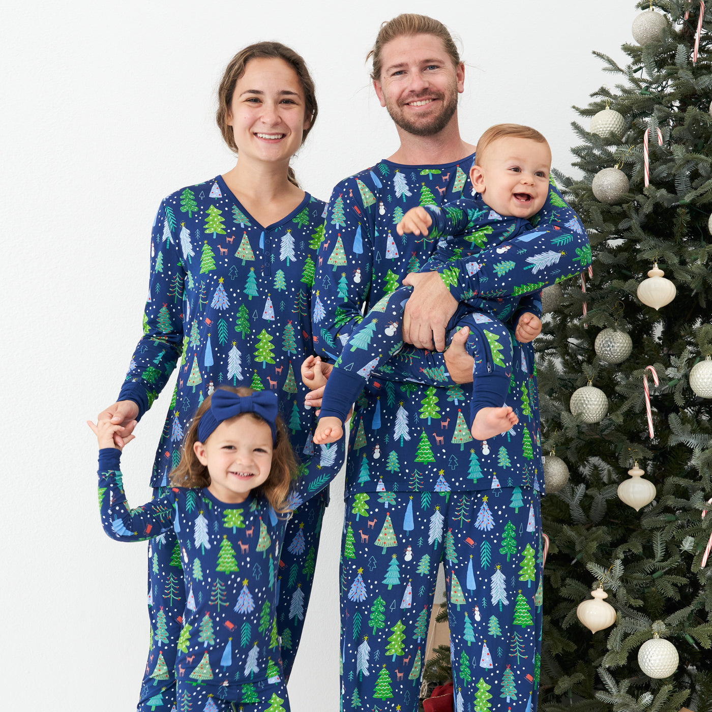 Family of four posing together wearing matching pajamas. Dad is wearing men's Blue Merry and Bright pajama bottoms paired with a matching Blue Merry and Bright pajama top. Mom is wearing women's Blue Merry and Bright pajama bottoms and a matching women's Blue Merry and Bright pajama top. Their kids are wearing a Blue Merry and Bright printed two piece pajama set paired with a Sapphire luxe bow headband and her brother is wearing a Blue Merry and Bright zippy