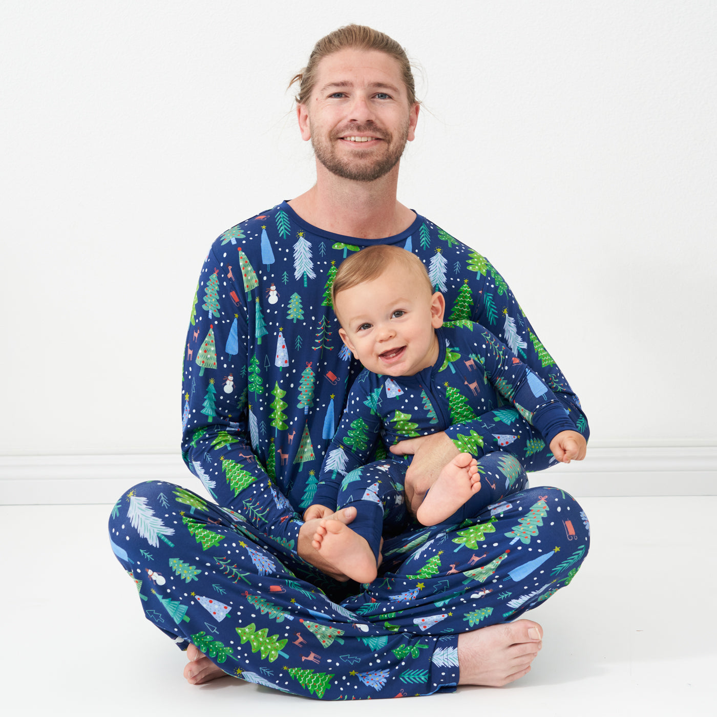 Father and son sitting together wearing matching Blue Merry and Bright pajamas. Dad is wearing Blue Merry and Bright printed men's pajama top and matching men's pajama bottoms and his son is wearing a Blue Merry and Bright printed zippy