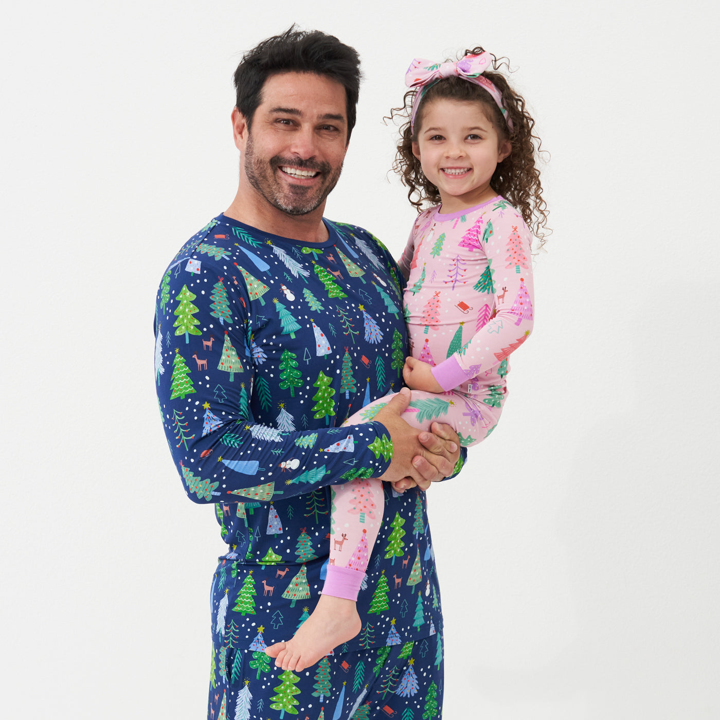 Father holding up his daughter. Dad is wearing Blue Merry and Bright printed pajama bottoms and a matching men's Blue Merry and Bright pajama top. His daughter is wearing Pink Merry and Bright two piece pajama set paired with a matching Pink Merry and Bright printed luxe bow headband