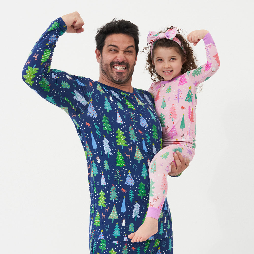 Alternative image of a father holding up his daughter. Dad is wearing Blue Merry and Bright printed pajama bottoms and a matching men's Blue Merry and Bright pajama top. His daughter is wearing Pink Merry and Bright two piece pajama set paired with a matching Pink Merry and Bright printed luxe bow headband