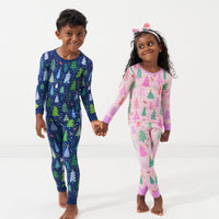 Two children holding hands wearing coordinating Pink and Blue Merry and Bright two piece pajama sets. The little girl has a her pjs paired with a matching Pink Merry and Bright luxe bow headband