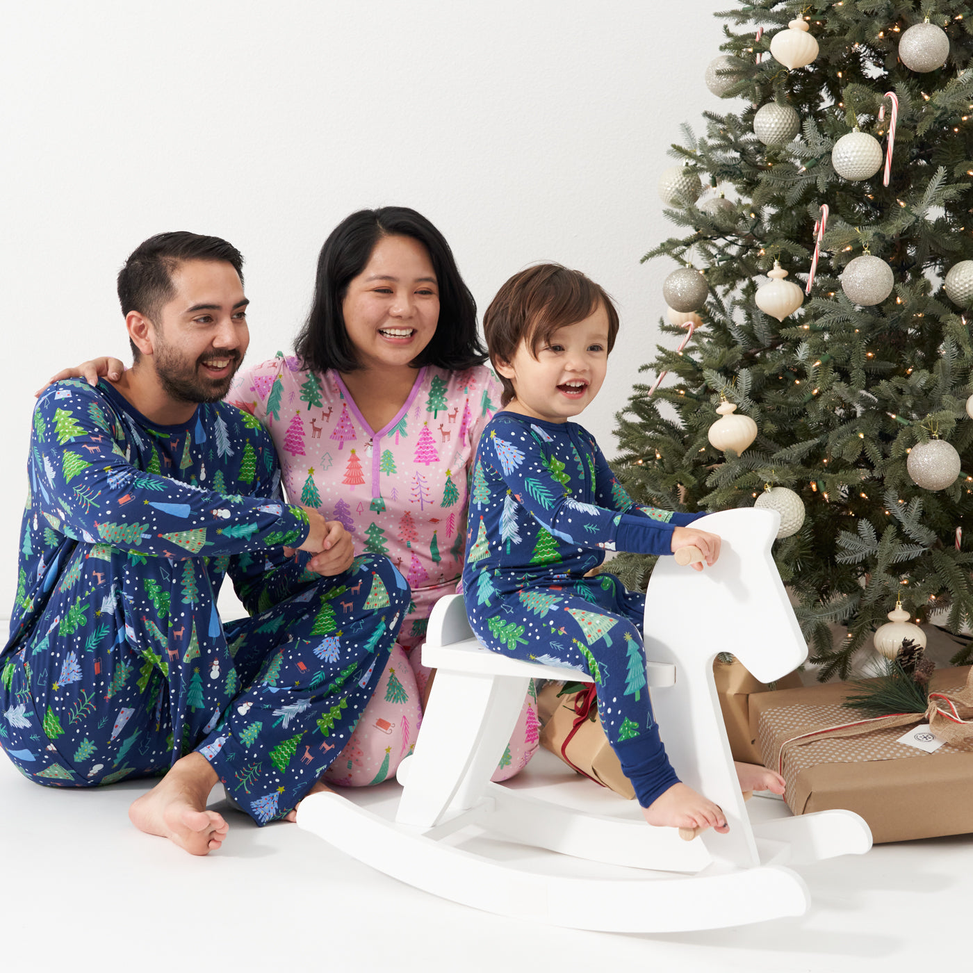 Family of three sitting around a Christmas tree. Dad is wearing Blue Merry and Bright printed men's pajama top and matching men's pajama bottoms. Mom is wearing Pink Merry and Bright women's pajama top paired with matching women's pajama bottoms. Their child is matching wearing a Blue Merry and Bright printed zippy