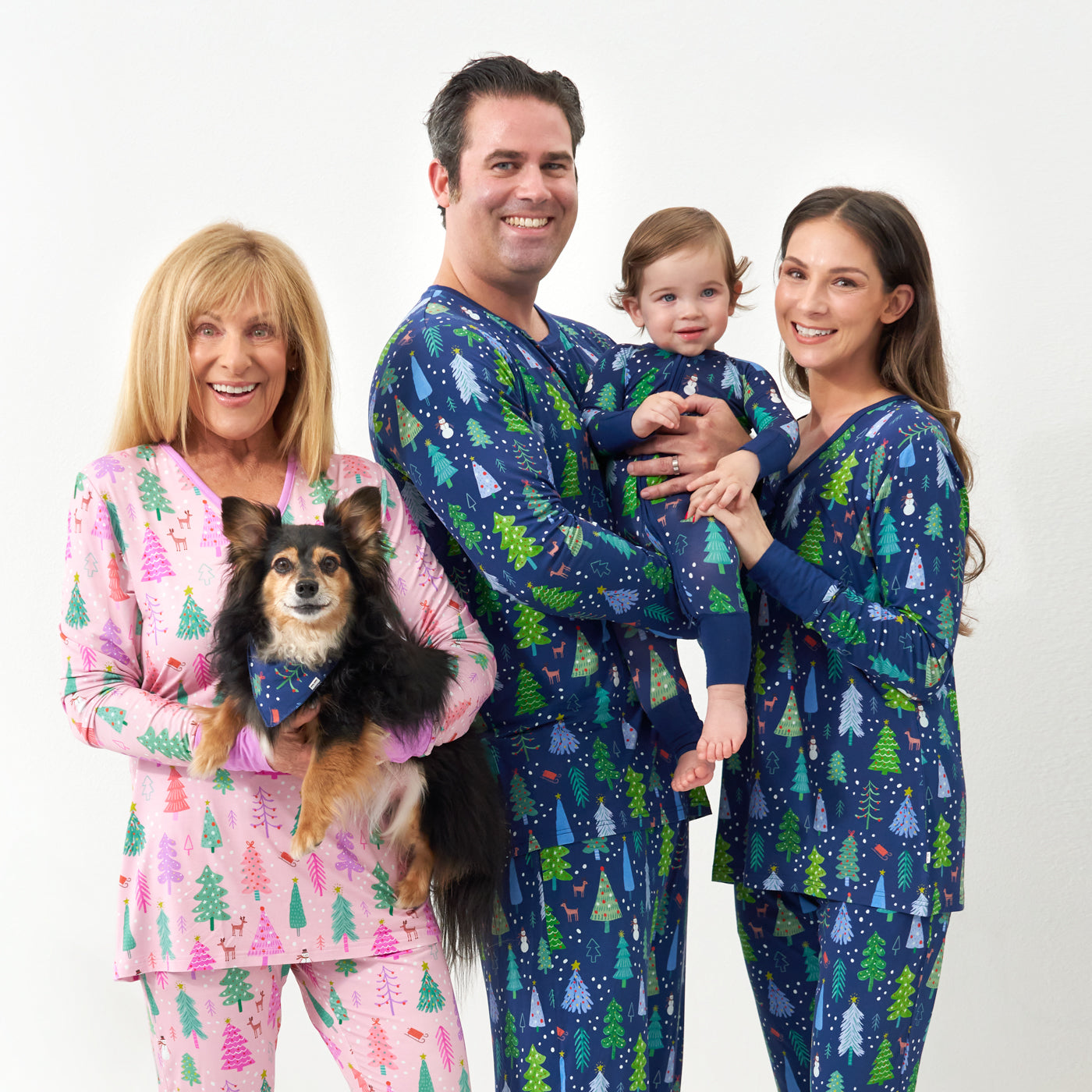 Family of four standing together with their dog. Grandma is wearing a women's Pink Merry and Bright women's pajama top and bottoms holding a Small dog wearing a Blue Merry and Bright pet bandana. Mom and Dad are wearing Blue Merry and Bright printed matching pajama tops and bottoms in men's and women's styles. Their child is matching wearing a Blue Merry and Bright zippy