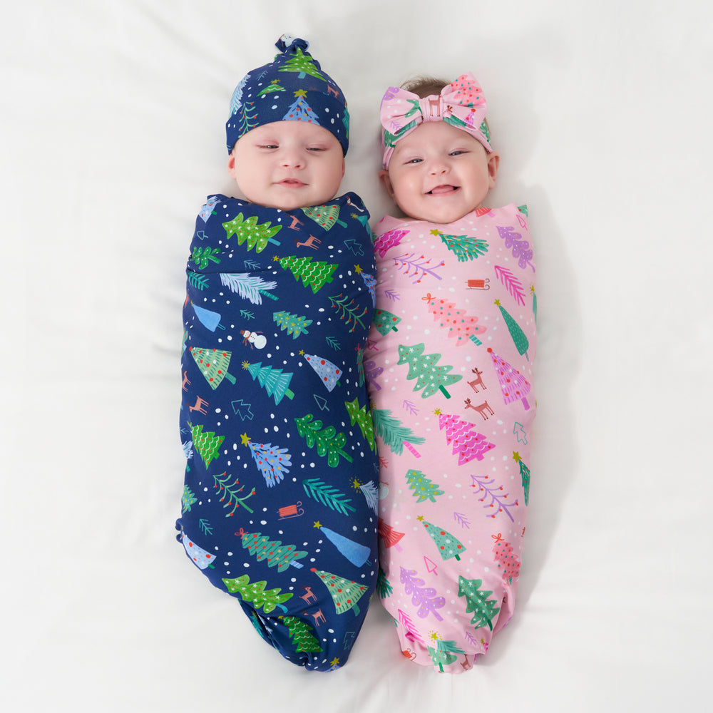 Two children laying on a bed swaddled in coordinating Blue Merry and Bright swaddle and hat set and a Pink Merry and Bright swaddle and luxe bow headband set