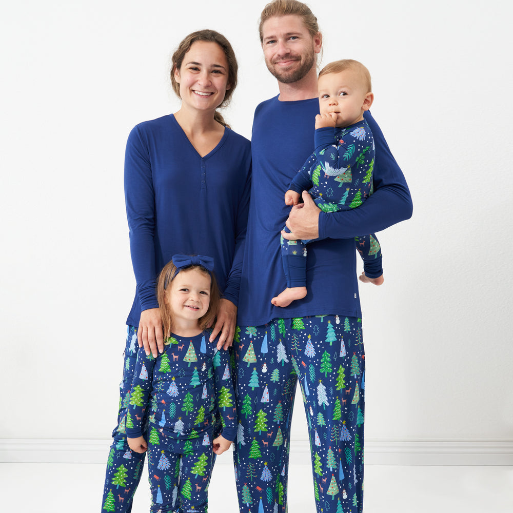 Family of four posing together wearing matching pajamas. Dad is wearing men's Blue Merry and Bright pajama bottoms paired with a solid Sapphire long sleeve pajama top. Mom is wearing women's Blue Merry and Bright pajama bottoms and a solid women's Sapphire long sleeve pajama top. Their kids are wearing a Blue Merry and Bright printed two piece pajama set paired with a Sapphire luxe bow headband and her brother is wearing a Blue Merry and Bright zippy