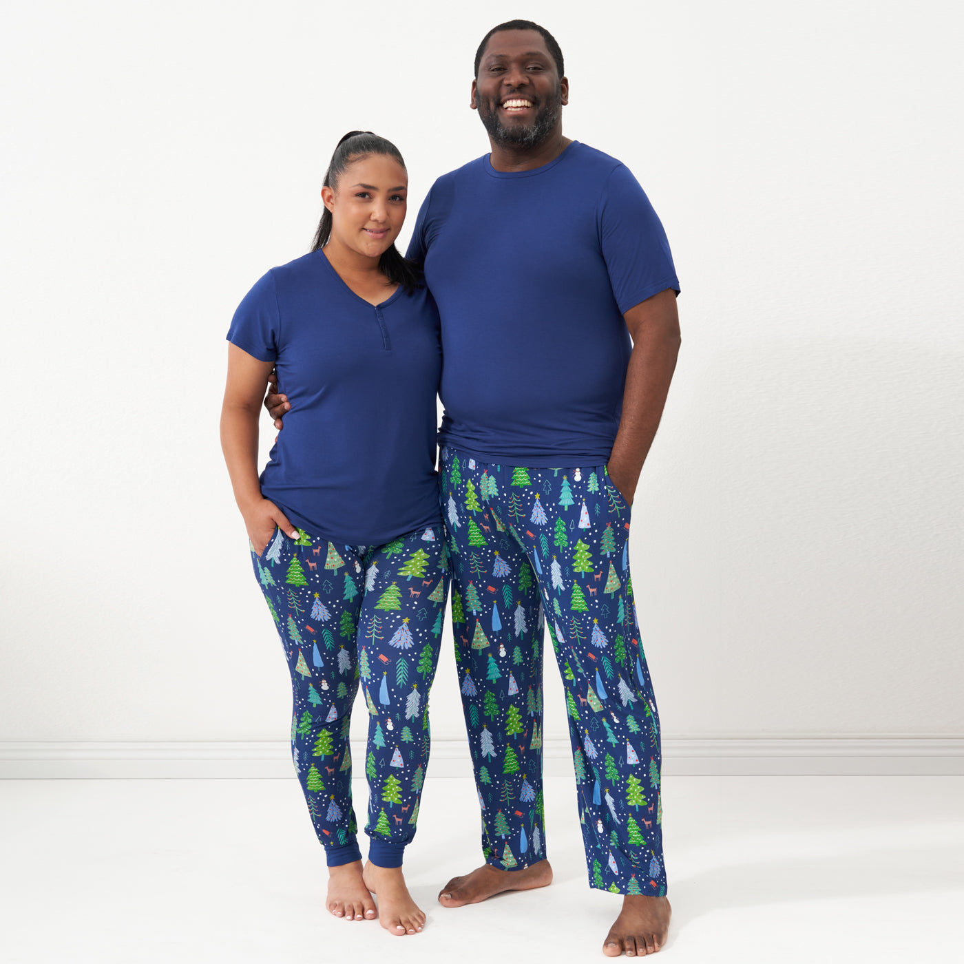 Couple posing together wearing matching pajamas. Man is wearing men's Blue Merry and Bright pajama bottoms paired with a solid Sapphire short sleeve pajama top. Woman is wearing women's Blue Merry and Bright pajama bottoms and a solid women's Sapphire short sleeve pajama top