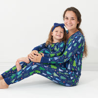 Mom and daughter posing together wearing matching Blue Merry and Bright pajamas. Mom is wearing women's Blue Merry and Bright printed pajama top and matching women's pajama bottoms. Daughter is wearing a Blue Merry and Bright two piece pajama set paired with a matching Sapphire luxe bow headband