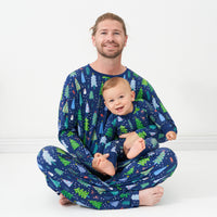 Father and son sitting together wearing matching Blue Merry and Bright pajamas. Dad is wearing Blue Merry and Bright printed men's pajama top and matching men's pajama bottoms and his son is wearing a Blue Merry and Bright printed zippy