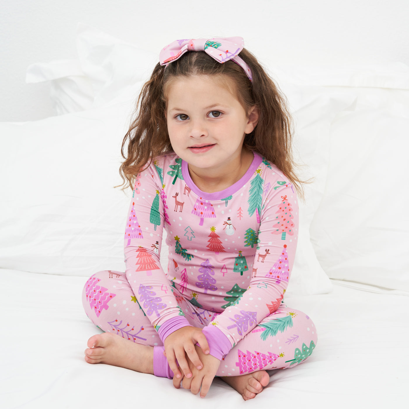 Child sitting wearing a Pink Merry and Bright two piece pajama set paired with a matching luxe bow headband