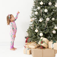 Child standing before a Christmas tree wearing a Pink Merry and Bright printed zippy paired with a matching Pink Merry and Bright luxe bow headband