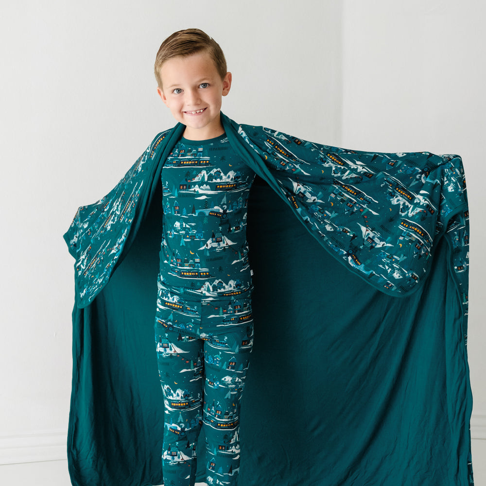 Alternate image of a child holding out a Midnight Express printed cloud blanket showing the solid teal backing. He is wearing matching Midnight Express printed two piece pajama set