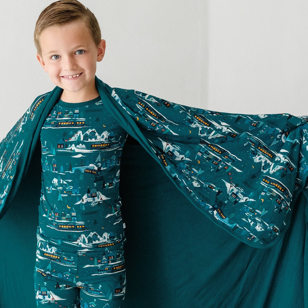 Close up image of a child holding out a Midnight Express printed cloud blanket showing the solid teal backing. He is wearing matching Midnight Express printed two piece pajama set